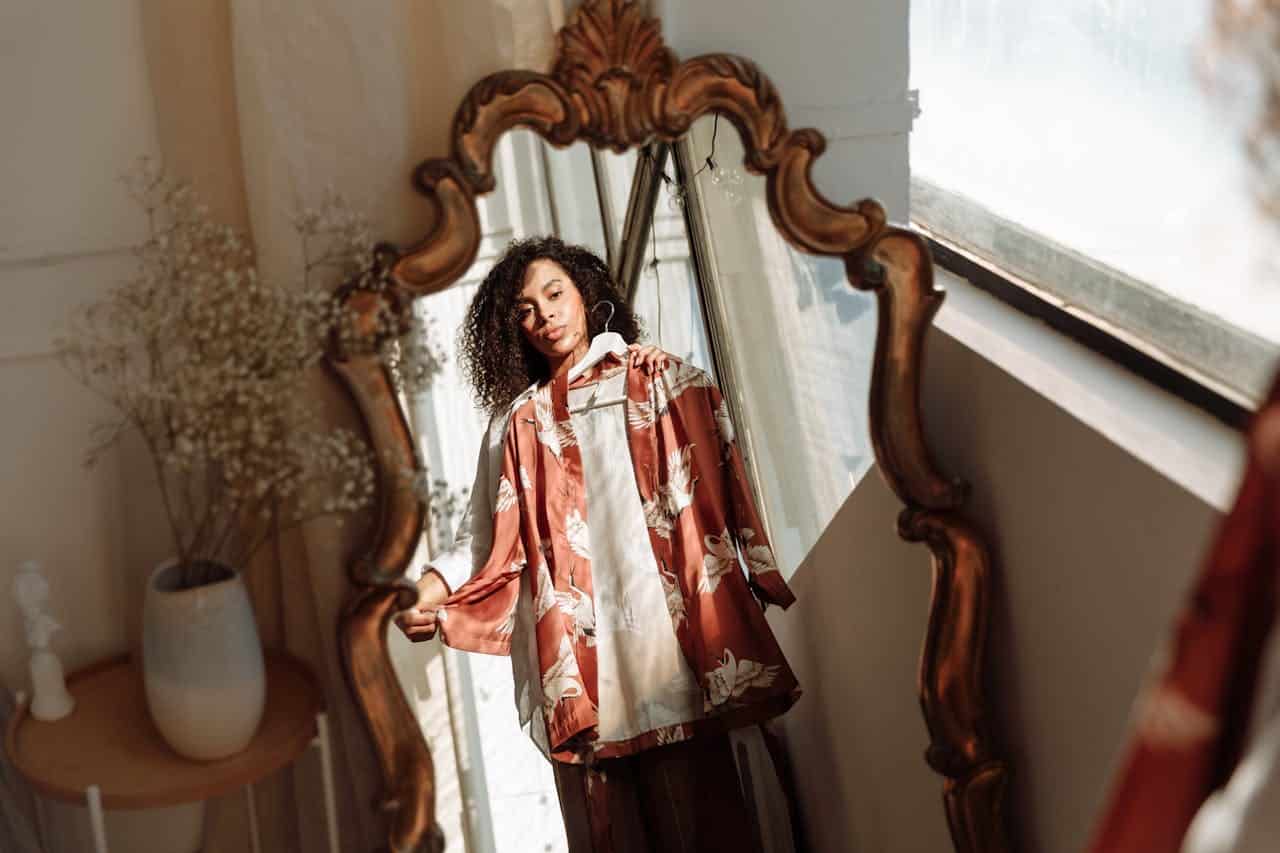 A person standing in front of a decorative mirror, holding a shirt, the reflection shows the individual and the environment, with sunlight streaming through a window, a glimpse of a vase and a wooden table in the background, the mirror's ornate frame adds a vintage touch to the scene