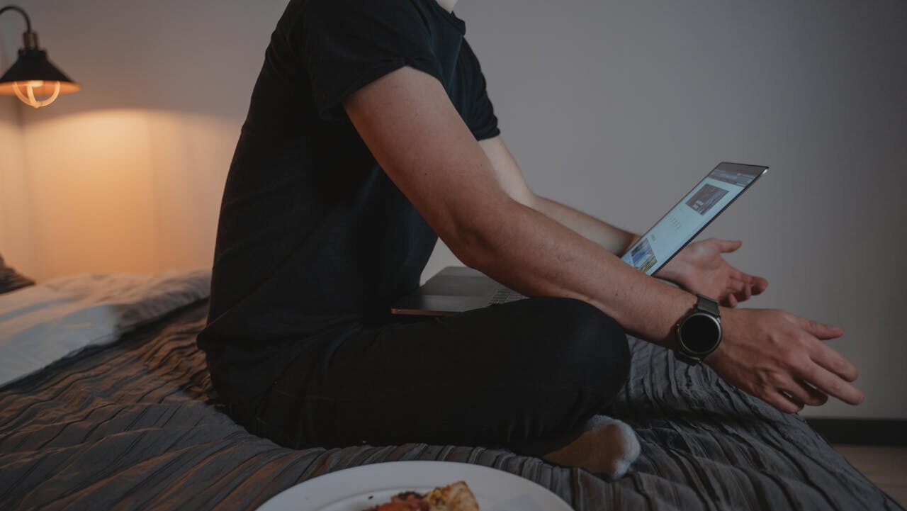 A person sitting on a bed with a laptop, wearing a black t-shirt and a smartwatch, eating pizza from a plate on the bed, looking at the screen, soft lighting creates a cozy and relaxed environment, engaging in a casual activity or online work from home scenario