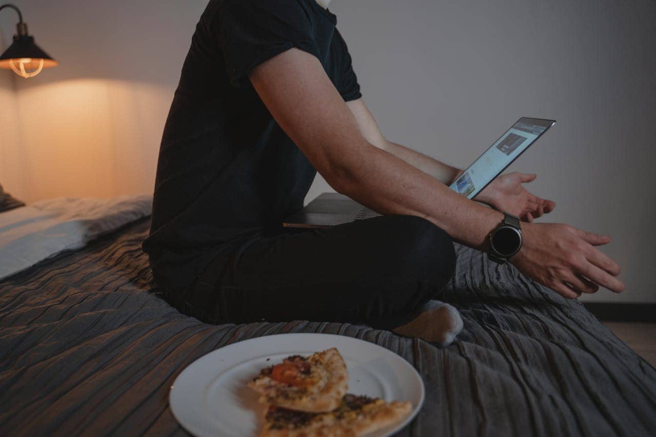 A person sitting on a bed with a laptop, wearing a black t-shirt and a smartwatch, eating pizza from a plate on the bed, looking at the screen, soft lighting creates a cozy and relaxed environment, engaging in a casual activity or online work from home scenario