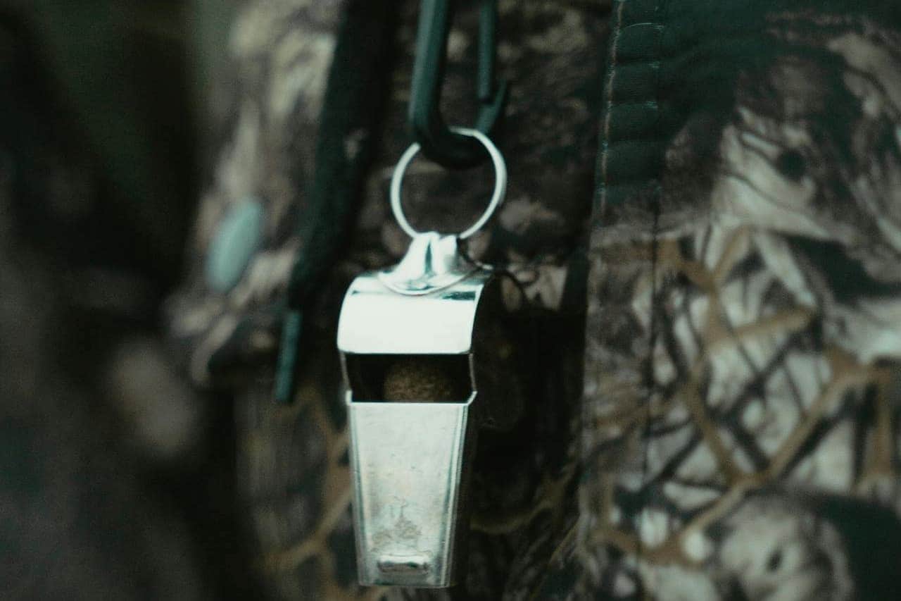 Metal whistle on keyring hanging from dark green strap against camouflage background, focused on reflective silver whistle body
