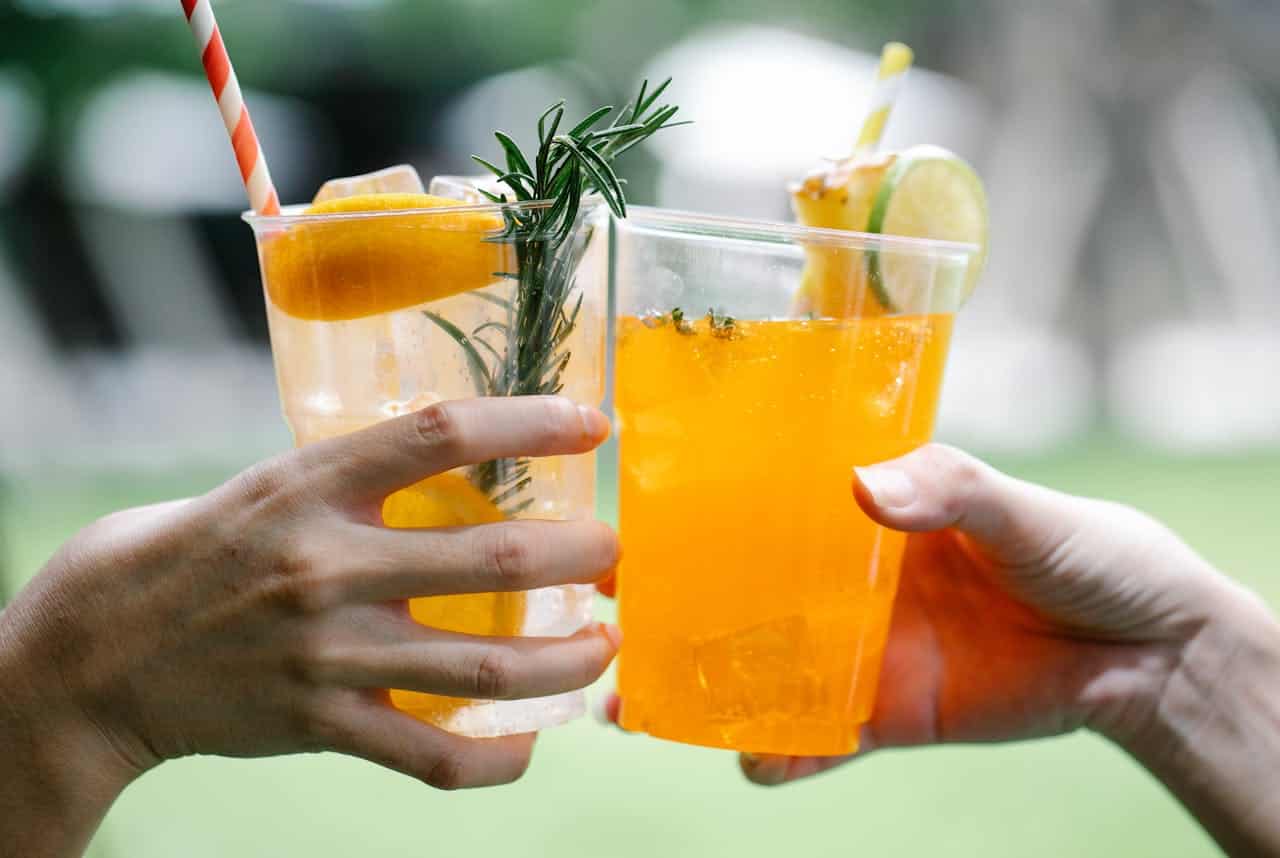 Two hands holding cups of citrus drinks, one with orange liquid, ice cubes, and a slice of orange and rosemary garnish, the other with orange liquid, ice, and a slice of lime