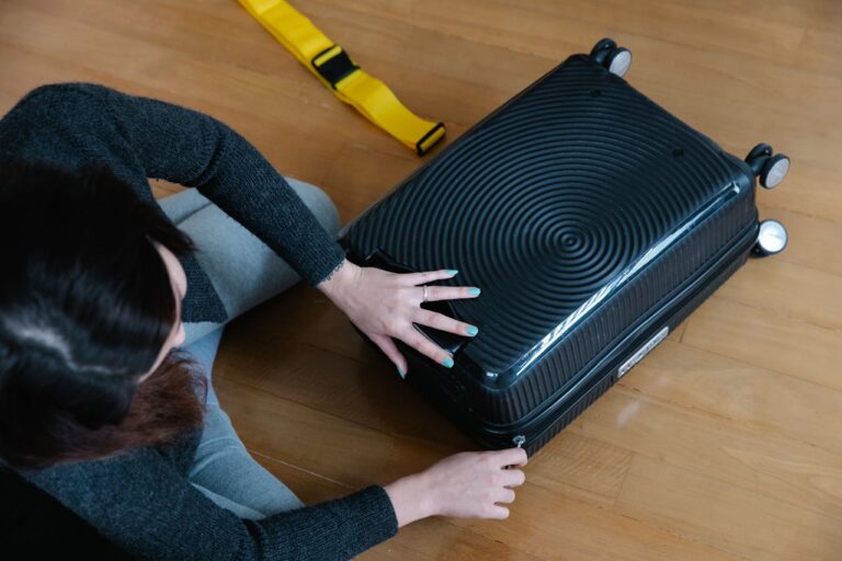 A person adjusting a black suitcase with a yellow strap on a wooden floor, preparing for travel, the suitcase is opened, the person's hands are positioned near the zipper
