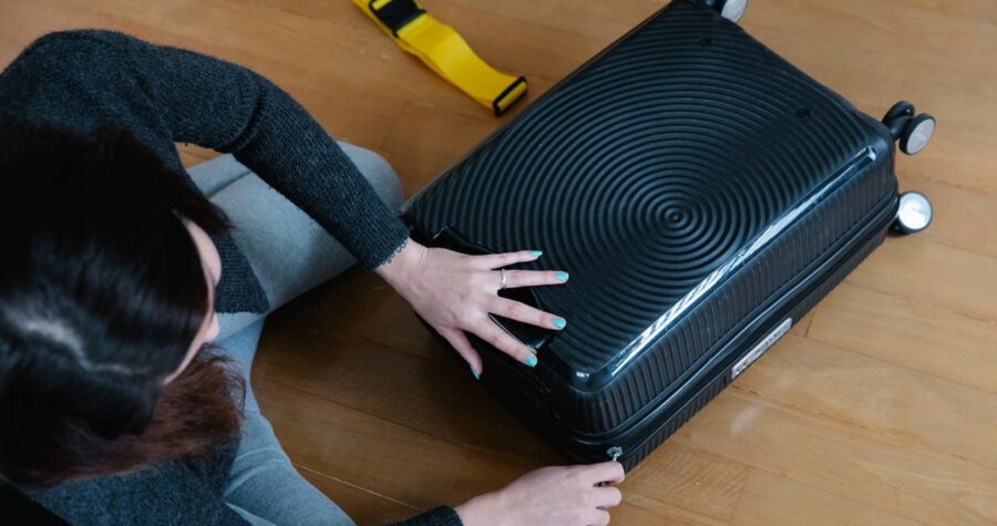 A person adjusting a black suitcase with a yellow strap on a wooden floor, preparing for travel, the suitcase is opened, the person's hands are positioned near the zipper
