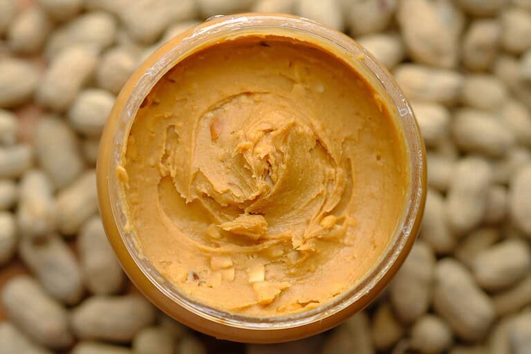 The image shows a jar of peanut butter, the creamy texture of the spread is visible inside the jar, peanut bits are scattered throughout the spread, peanuts are visible in the background
