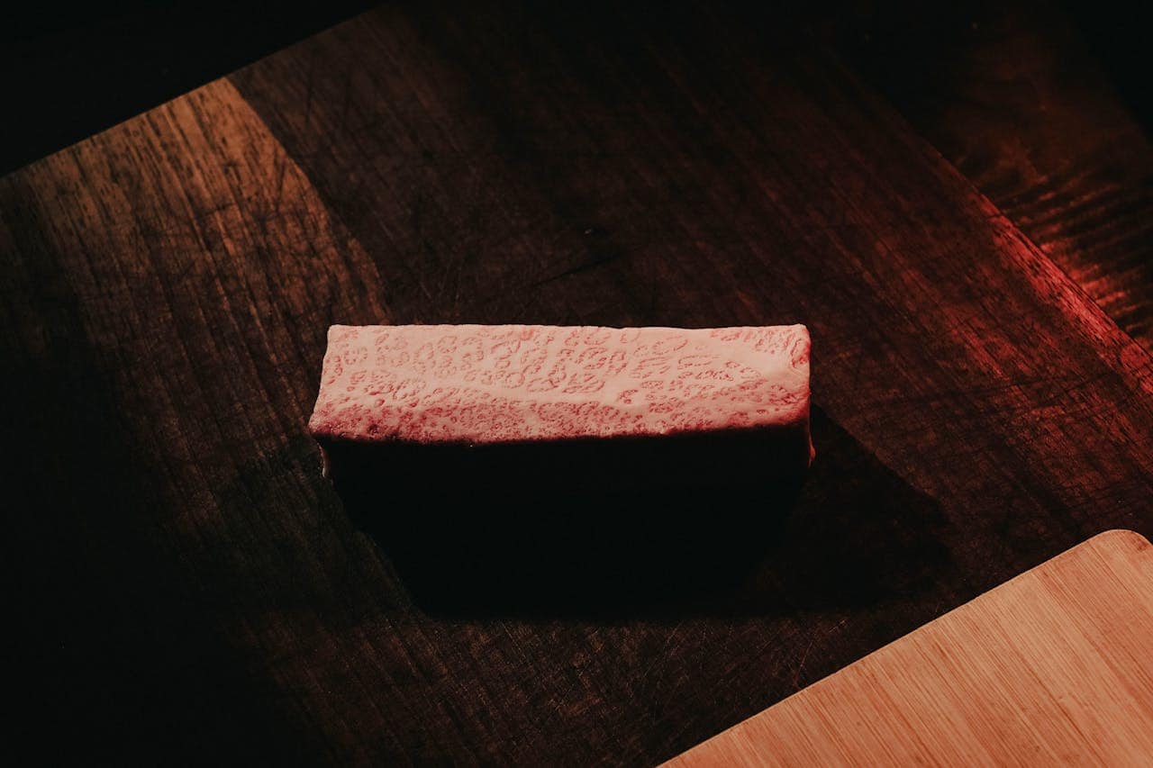 A piece of Wagyu beef is displayed on a wooden surface, illuminated by warm lighting that highlights its marbled texture