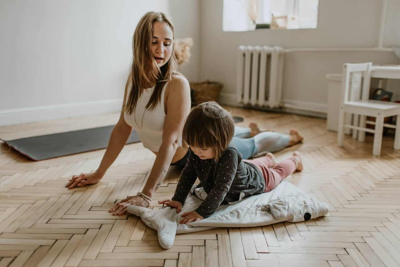 A woman practicing yoga with a child, both lying on the floor, child playing with a pillow, bright indoor setting, wooden floor, mother and child spending quality time together, exercise and bonding