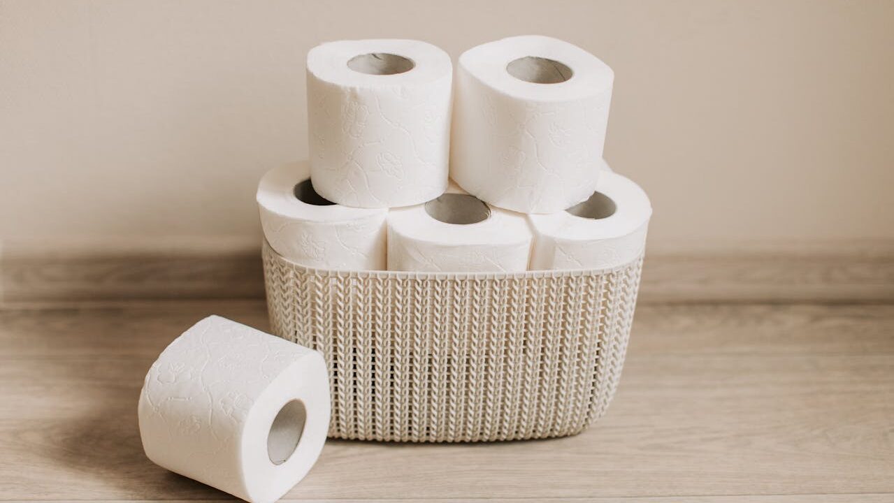 A stack of toilet paper rolls in a woven basket, several rolls are stacked neatly, some are positioned sideways, the rolls are white with a textured design, the basket is light in color
