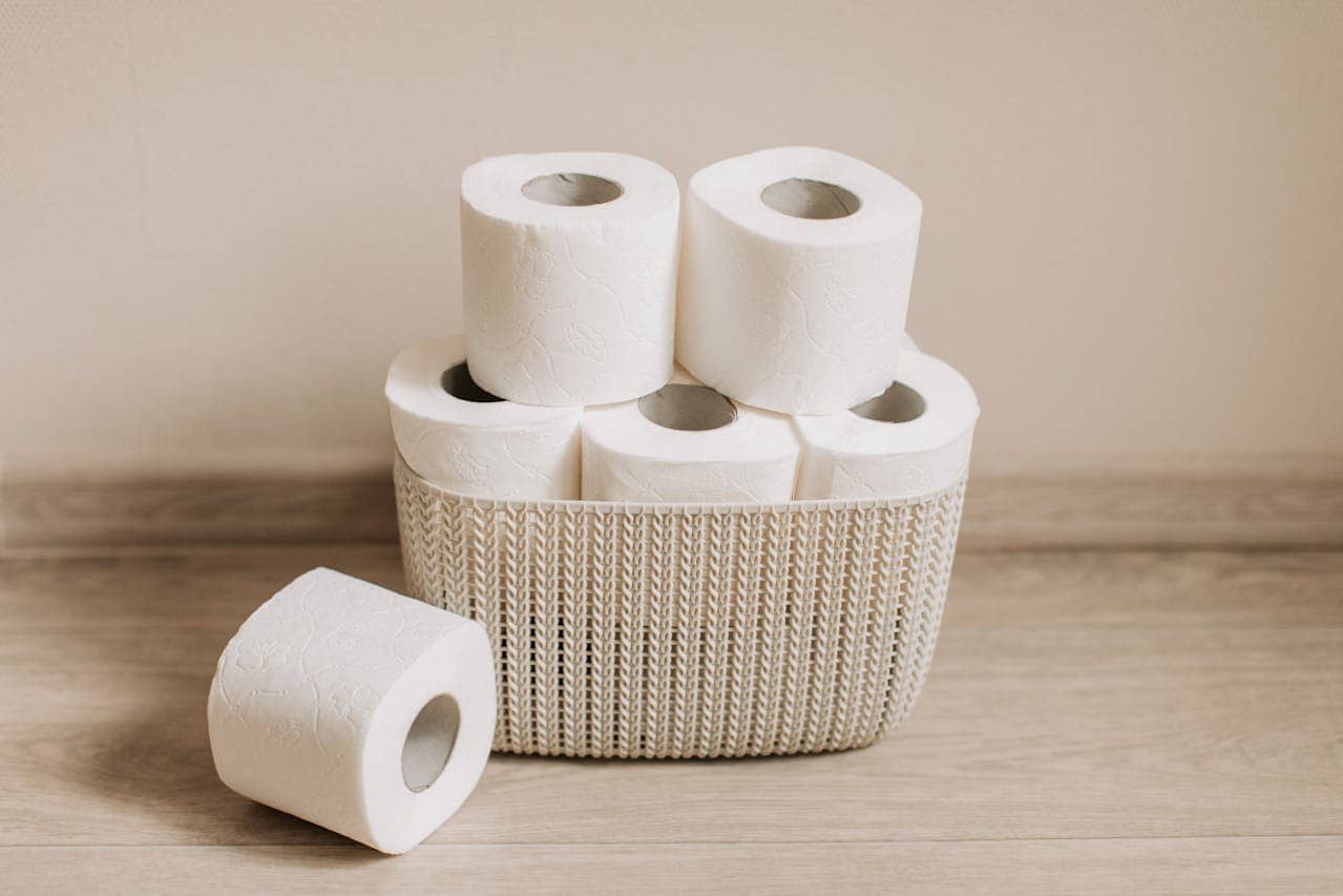 A stack of toilet paper rolls in a woven basket, several rolls are stacked neatly, some are positioned sideways, the rolls are white with a textured design, the basket is light in color