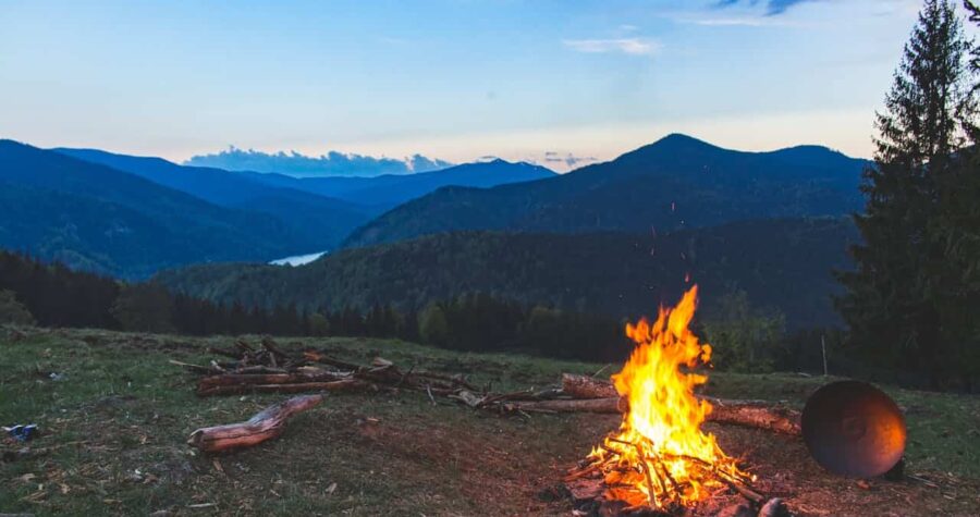 Campfire burning in the wilderness, mountainous landscape in the background, serene outdoor setting, warm firelight at dusk, peaceful campsite scene, wood logs stacked nearby, clear skies, nature and adventure, camping at sunset, fire for warmth and cooking