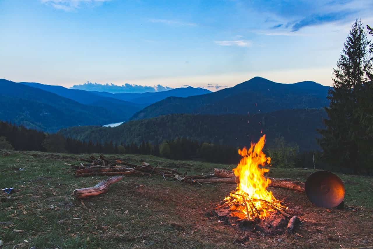 Campfire burning in the wilderness, mountainous landscape in the background, serene outdoor setting, warm firelight at dusk, peaceful campsite scene, wood logs stacked nearby, clear skies, nature and adventure, camping at sunset, fire for warmth and cooking