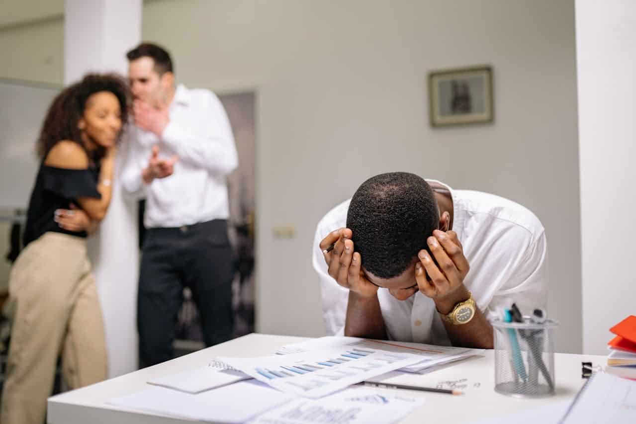 A man looking stressed and overwhelmed, sitting at a desk with papers and graphs, while two colleagues in the background whisper and laugh, symbolizing peer pressure