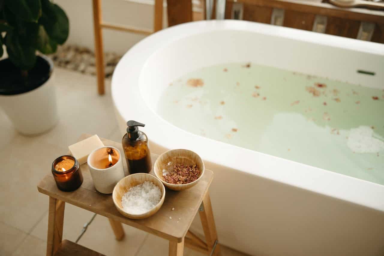 A wooden stool with candles, soap, essential oil, bath salts, and dried flowers placed beside a bath filled with water and rose petals, a relaxing, soothing spa setup, a peaceful atmosphere with a calm and serene bathroom design