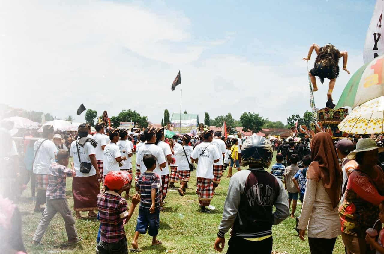 A vibrant outdoor festival, people gathering and walking, individuals wearing traditional attire, a large puppet being carried above the crowd, children and adults enjoying the event, banners and umbrellas in the background