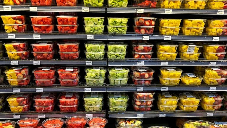 Supermarket display of fresh produce in plastic containers, arranged in rows on refrigerated shelves, featuring red tomatoes, yellow peppers, and green vegetables in transparent packaging
