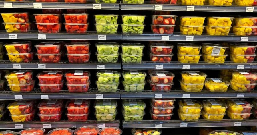Supermarket display of fresh produce in plastic containers, arranged in rows on refrigerated shelves, featuring red tomatoes, yellow peppers, and green vegetables in transparent packaging