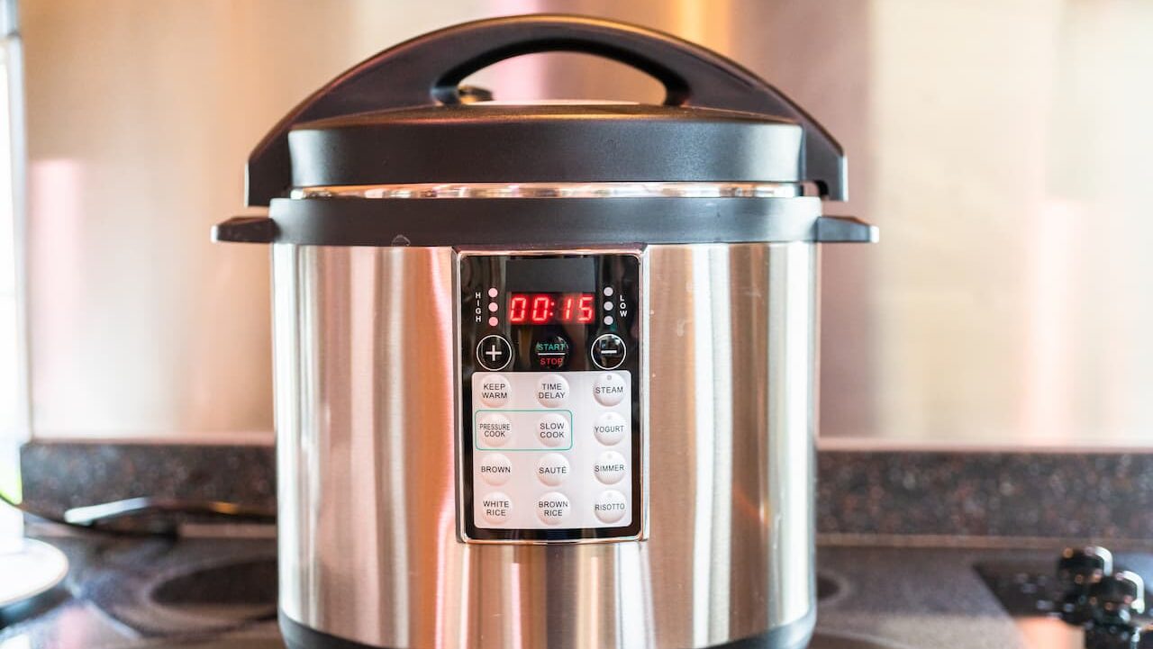 Stainless steel pressure cooker, digital display showing 15 minutes, multiple cooking functions, black lid with handle, placed on a stovetop, kitchen setting, modern appliance