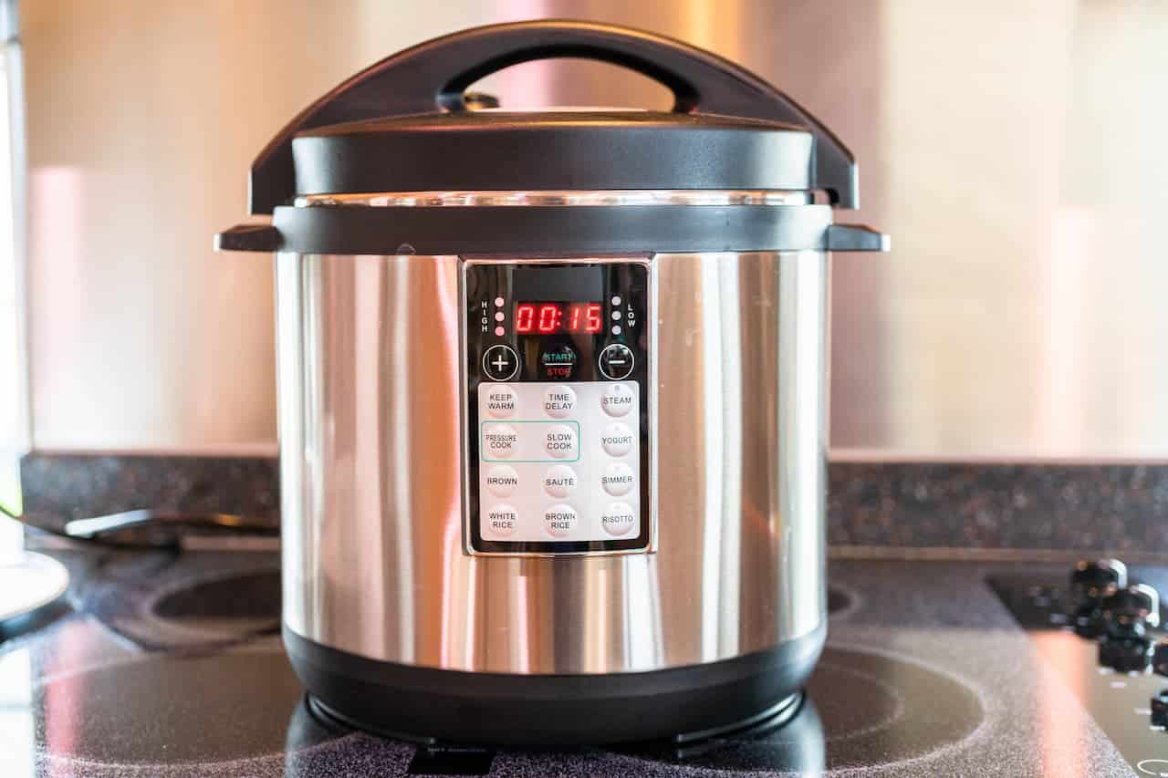 Stainless steel pressure cooker, digital display showing 15 minutes, multiple cooking functions, black lid with handle, placed on a stovetop, kitchen setting, modern appliance