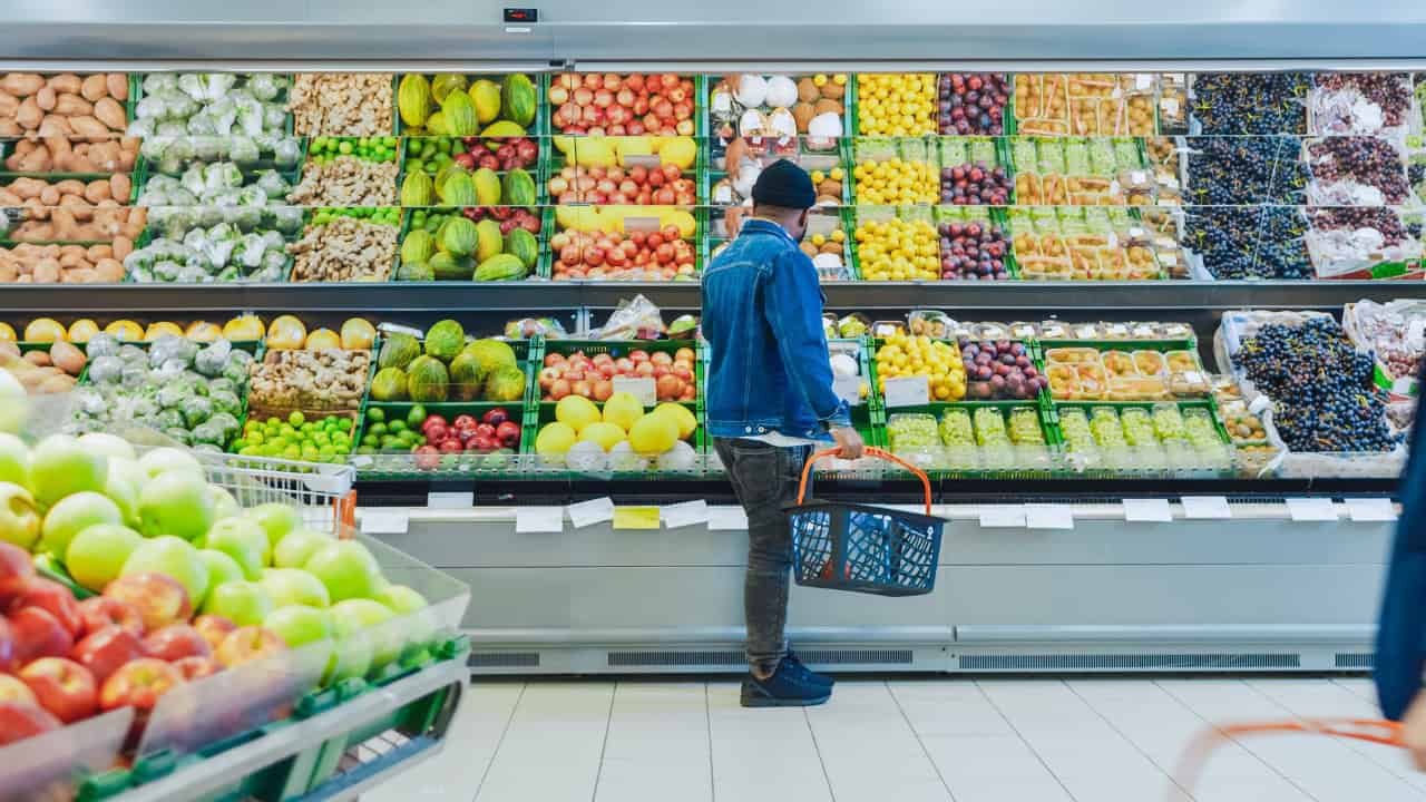 Person shopping for fresh produce in a grocery store, standing in front of a colorful selection of fruits, holding a shopping basket