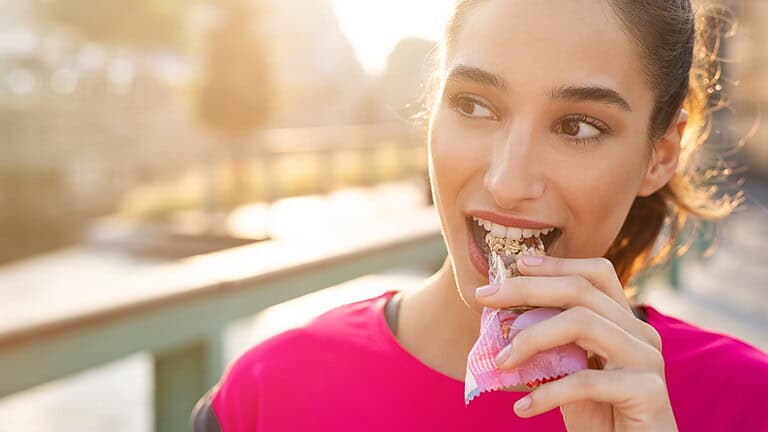 Woman in a pink sports shirt, smiling while biting into a granola bar, outdoor setting with sunlight, blurred background of a park or pathway