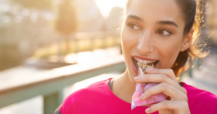 Woman in a pink sports shirt, smiling while biting into a granola bar, outdoor setting with sunlight, blurred background of a park or pathway
