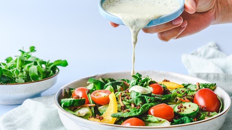 A fresh salad with spinach, cherry tomatoes, and yellow bell peppers, a hand pouring creamy dressing from a small bowl, bright and clean background
