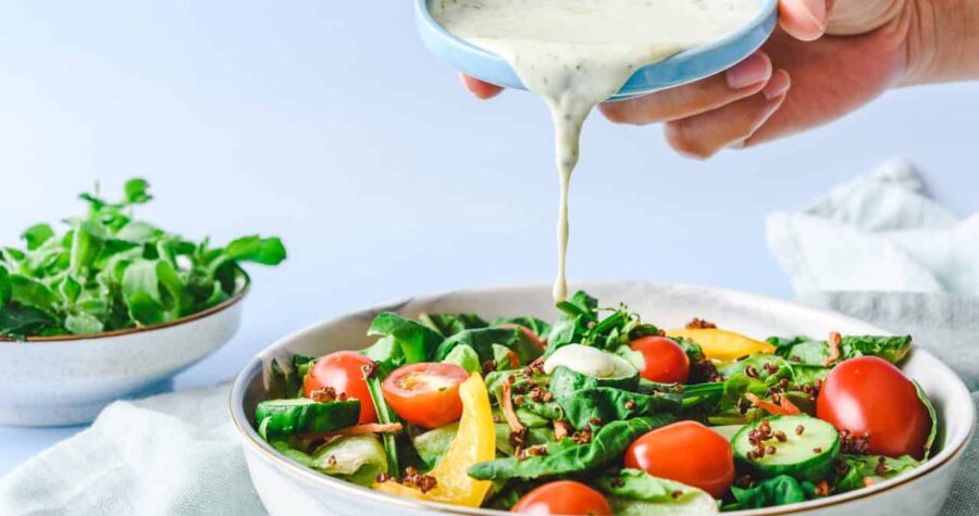 A fresh salad with spinach, cherry tomatoes, and yellow bell peppers, a hand pouring creamy dressing from a small bowl, bright and clean background