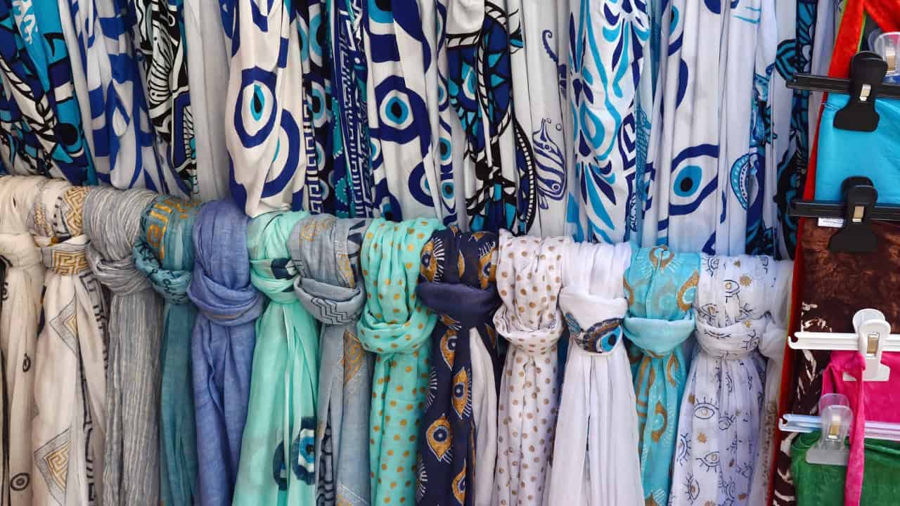 Collection of decorative scarves and textile trims in blue, turquoise, and cream colors hanging together. Features ornate patterns and beaded details. A blue square with white checkmark appears in top right corner