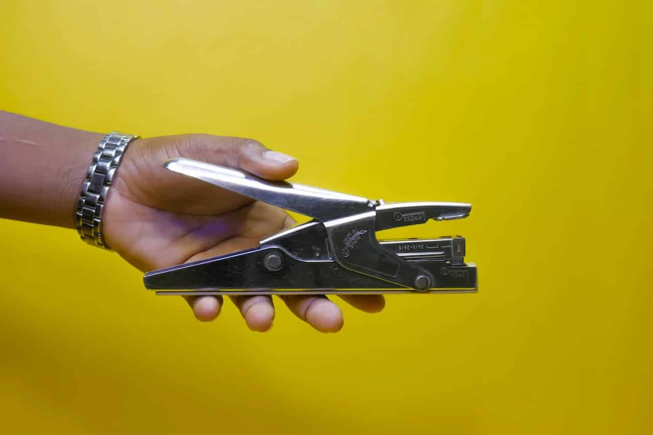Hand wearing metal watch holding manual can opener against bright yellow background, showing mechanical office tool