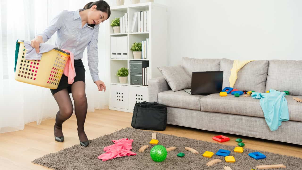 A woman in office attire, bending down to pick up toys and scattered objects, child’s play items on a rug, casual living room setting, sofa in the background