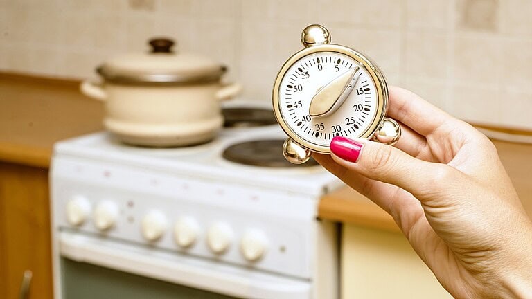 Hand with red-painted nails holding a kitchen timer set to approximately 35 minutes, stove and cooking pot in the background