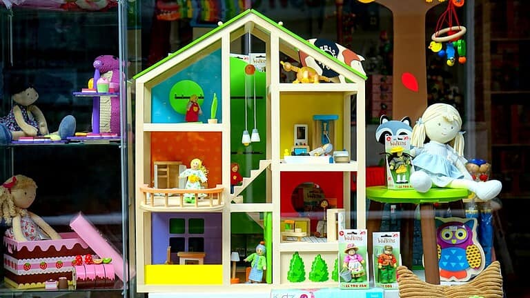 Colorful wooden dollhouse with miniature furniture and figurines, displayed in a toy store window, surrounded by plush dolls, packaged toy figurines, and decorative elements, vibrant and inviting children's playset