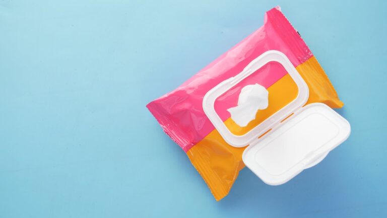Opened pack of wet wipes with a colorful pink and orange packaging, placed on a light blue surface, with one wipe partially pulled through the plastic dispenser lid
