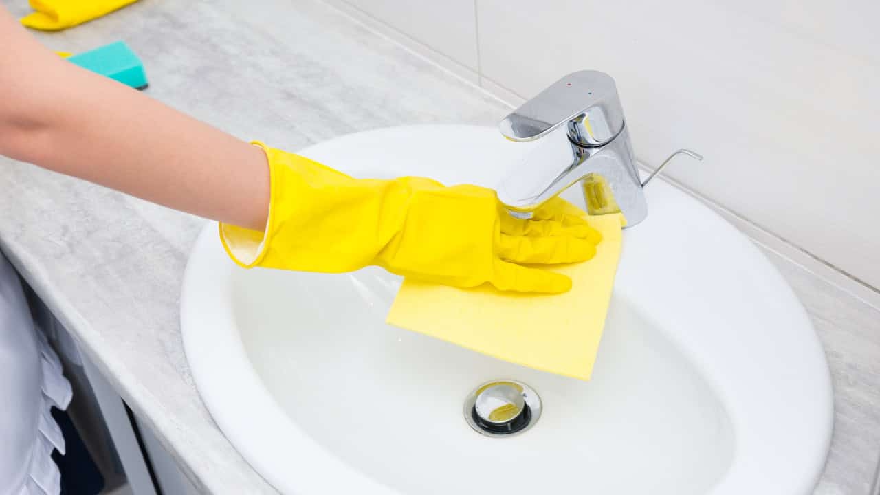Person wearing yellow rubber gloves, wiping a bathroom sink faucet, cleaning with a sponge, maintaining hygiene in the sink, household cleaning task