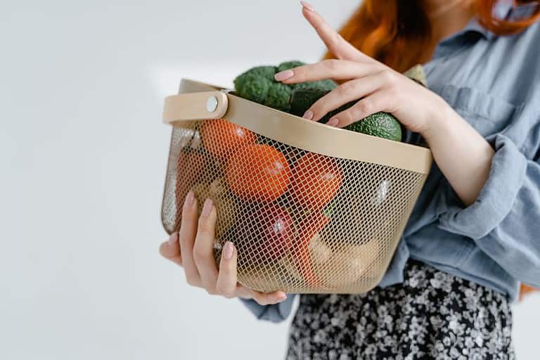 Woman holding a basket of fresh vegetables, including tomatoes, avocados, onions, and broccoli, wearing a blue shirt and floral skirt, healthy grocery shopping, fresh produce storage