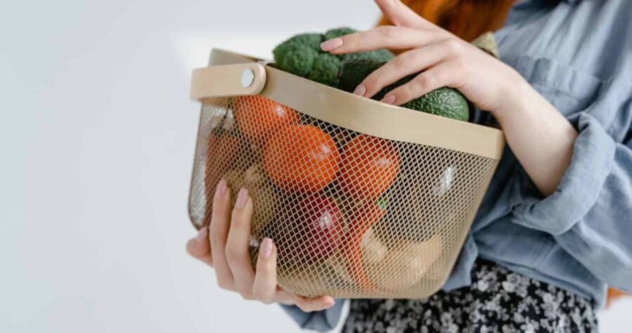 Woman holding a basket of fresh vegetables, including tomatoes, avocados, onions, and broccoli, wearing a blue shirt and floral skirt, healthy grocery shopping, fresh produce storage