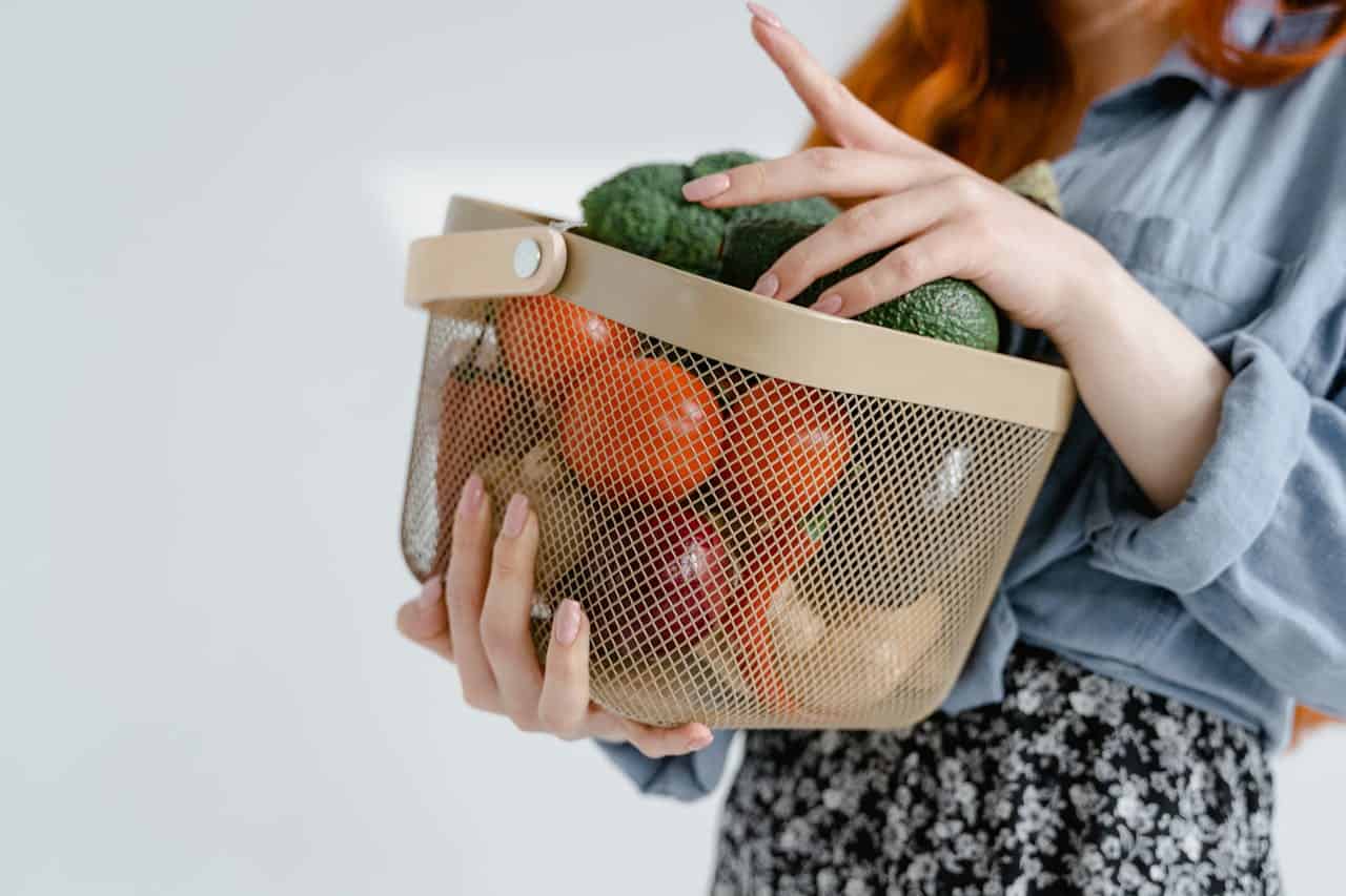 Woman holding a basket of fresh vegetables, including tomatoes, avocados, onions, and broccoli, wearing a blue shirt and floral skirt, healthy grocery shopping, fresh produce storage