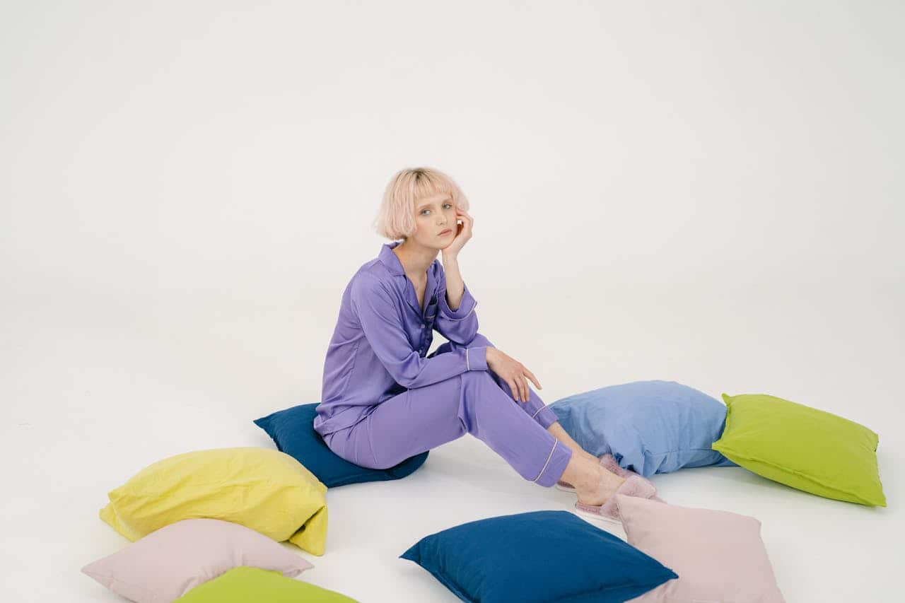 Person with short blonde hair wearing purple pajamas, sitting among colorful pillows, white background, contemplative pose