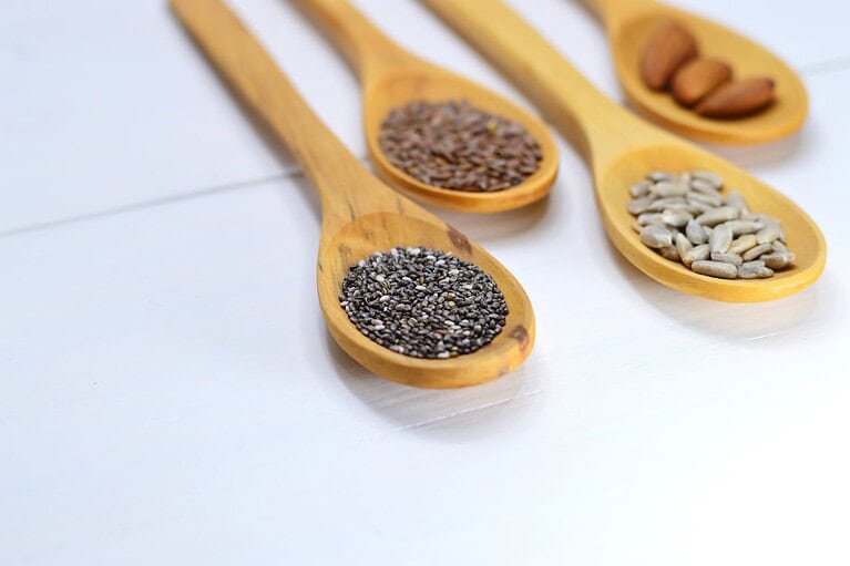 Wooden spoons filled with chia seeds, flaxseeds, sunflower seeds, and almonds, arranged on a white surface, healthy superfoods, natural ingredients, nutrition-rich seeds