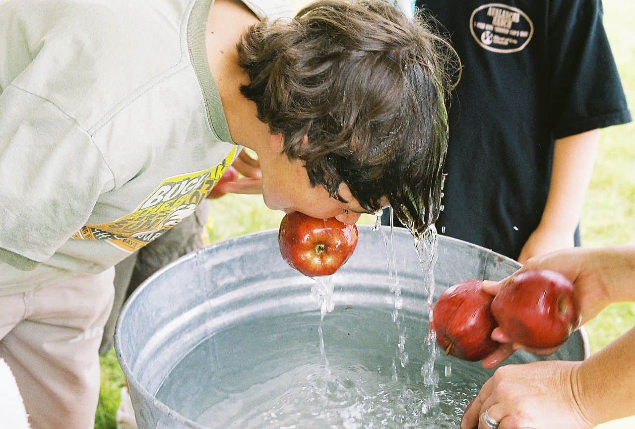A child participating in a traditional apple bobbing game, the child has an apple in their mouth, water splashes around the apple, other apples are held by hands outside the tub, a joyful outdoor activity, the child is drenched with water