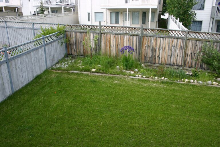 A well-maintained backyard with a grassy lawn, a wooden fence with a lattice top, flower beds along the fence with blooming purple flowers and green plants, bordered with small rocks, a quiet residential area in the background with a few houses visible