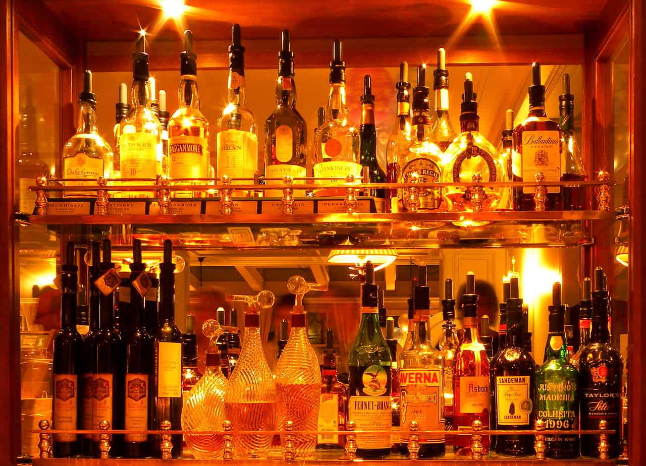 The image shows a well-lit bar shelf, showcasing an array of bottles of liquor, including whiskey, brandy, and other spirits, all neatly arranged on golden shelves with warm, glowing lights, creating a luxurious and inviting atmosphere