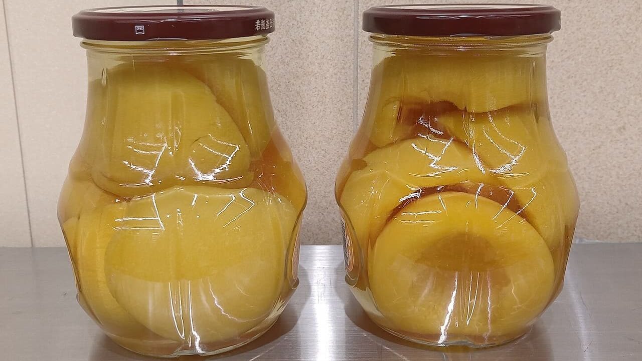 Two glass jars with brown lids containing preserved yellow peaches, in syrup against light background