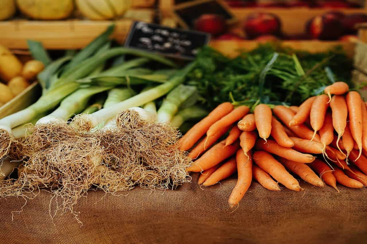 Fresh vegetables including leeks with visible roots, leafy herbs, and orange carrots on burlap fabric