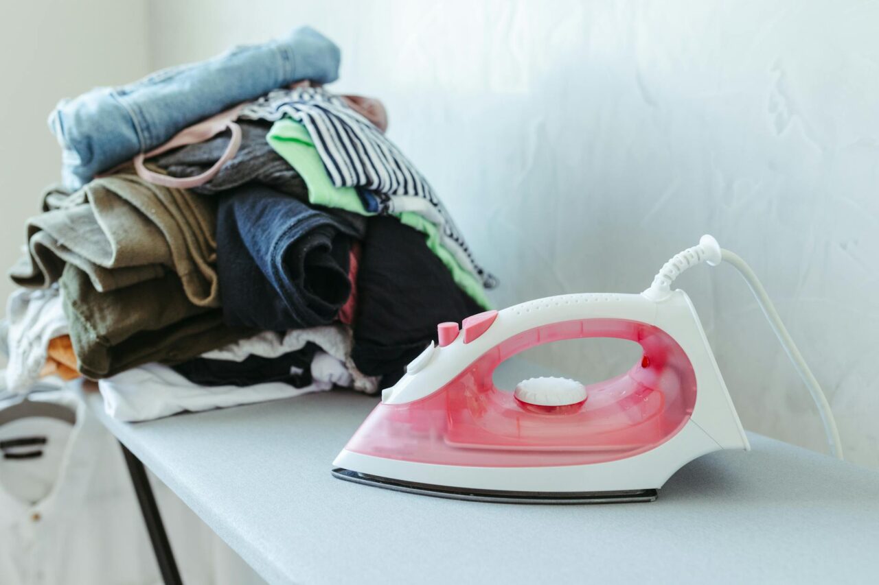A red iron placed beside a stack of colorful clothes on an ironing board indoors