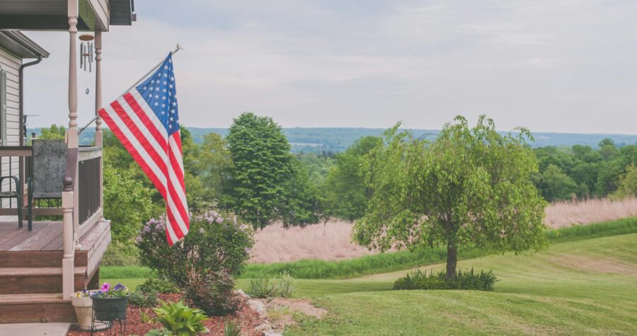 A serene suburban house porch displaying an American flag with a scenic background.