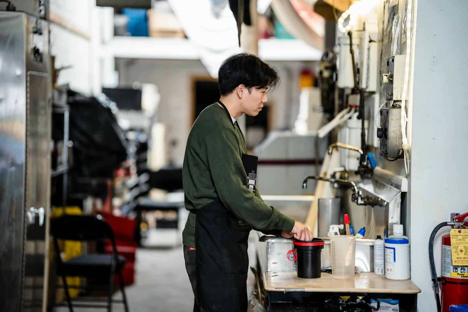 Asian man in green sweater mixing chemicals in an industrial workspace indoors, focused on work.