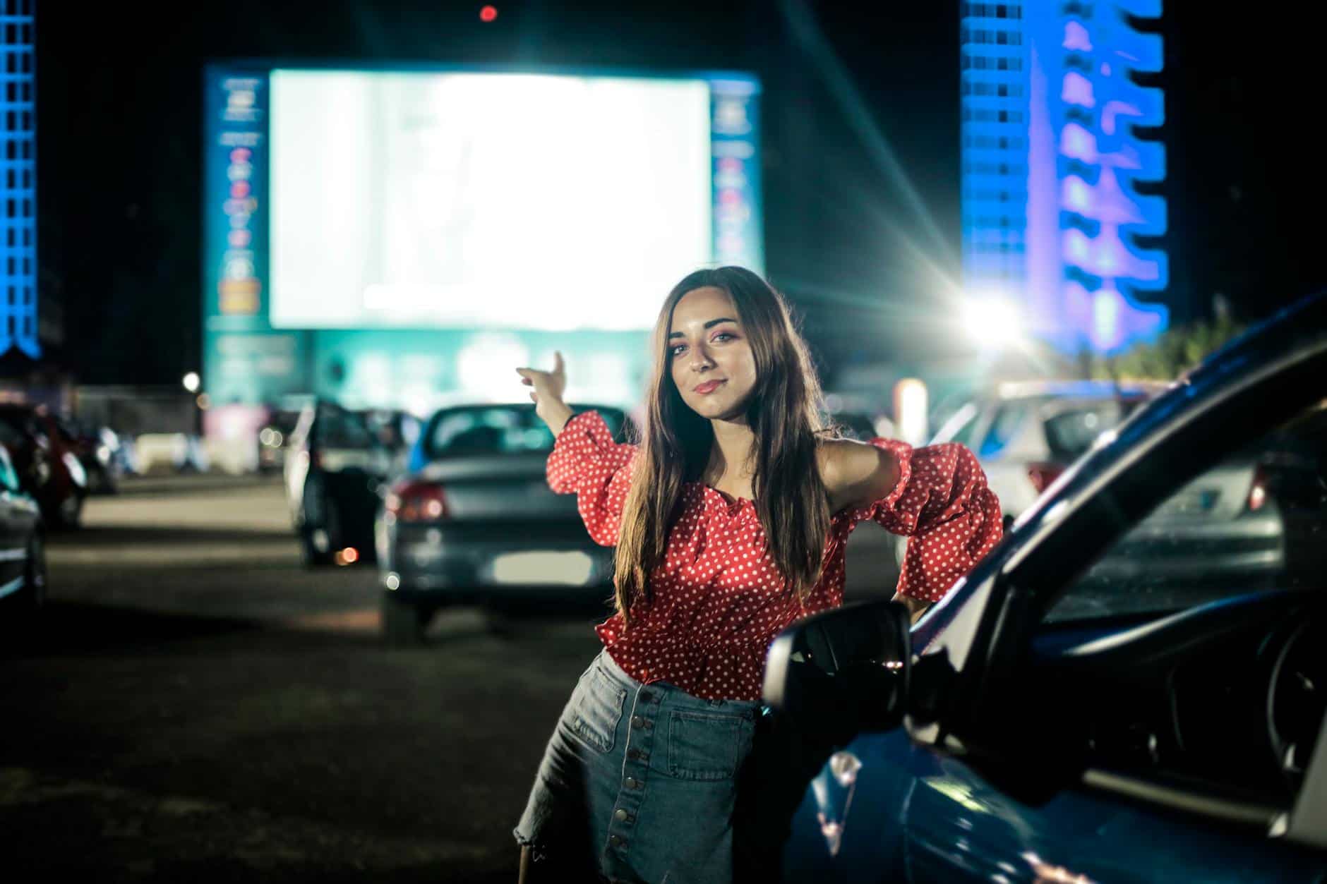 Cheerful female in trendy outfit looking at camera while standing near car on parking lot in evening time and showing hand on screen