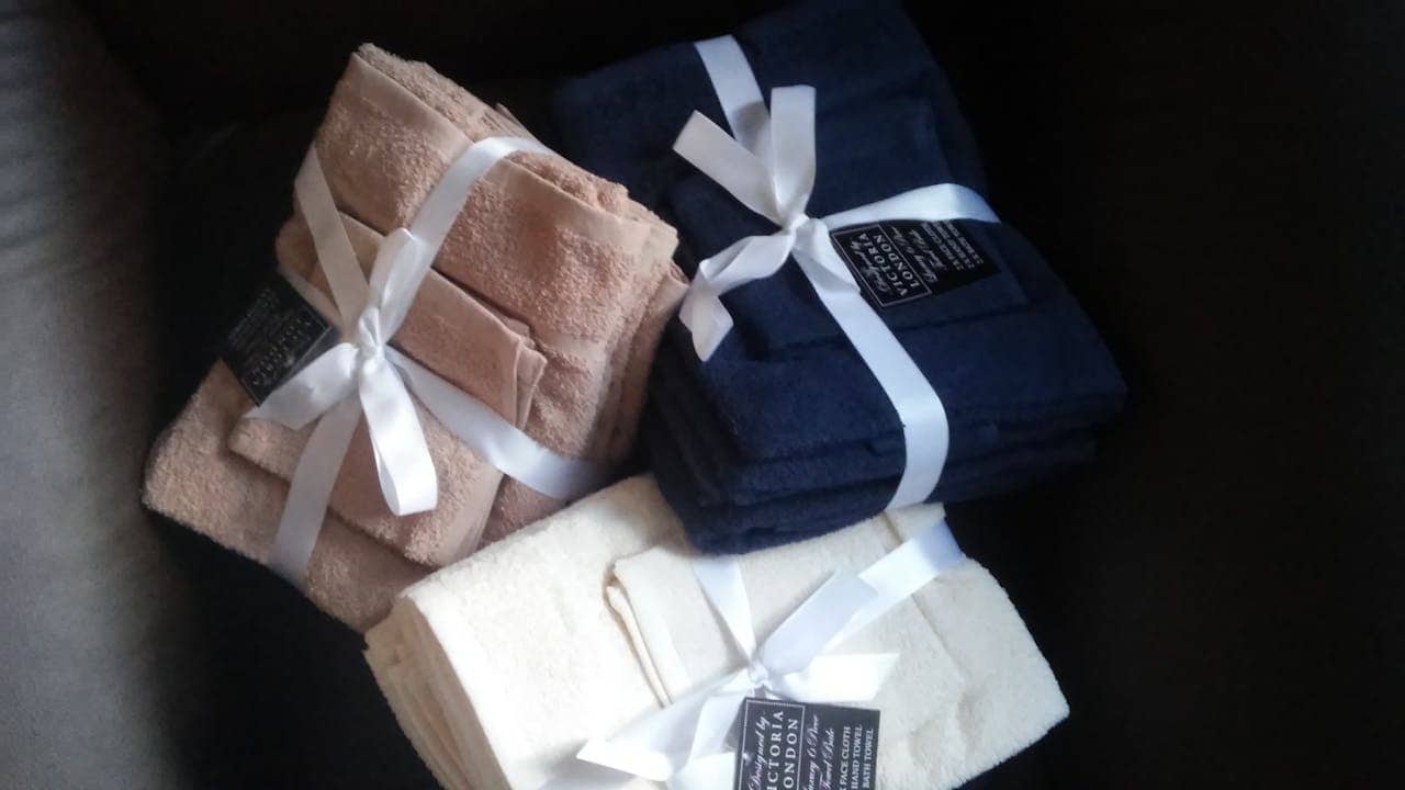 Three stacks of folded towels tied with white ribbon bows. Sets include blush pink, navy blue, and cream-colored towels, each with a black branded tag attached. Arranged on what appears to be a dark surface