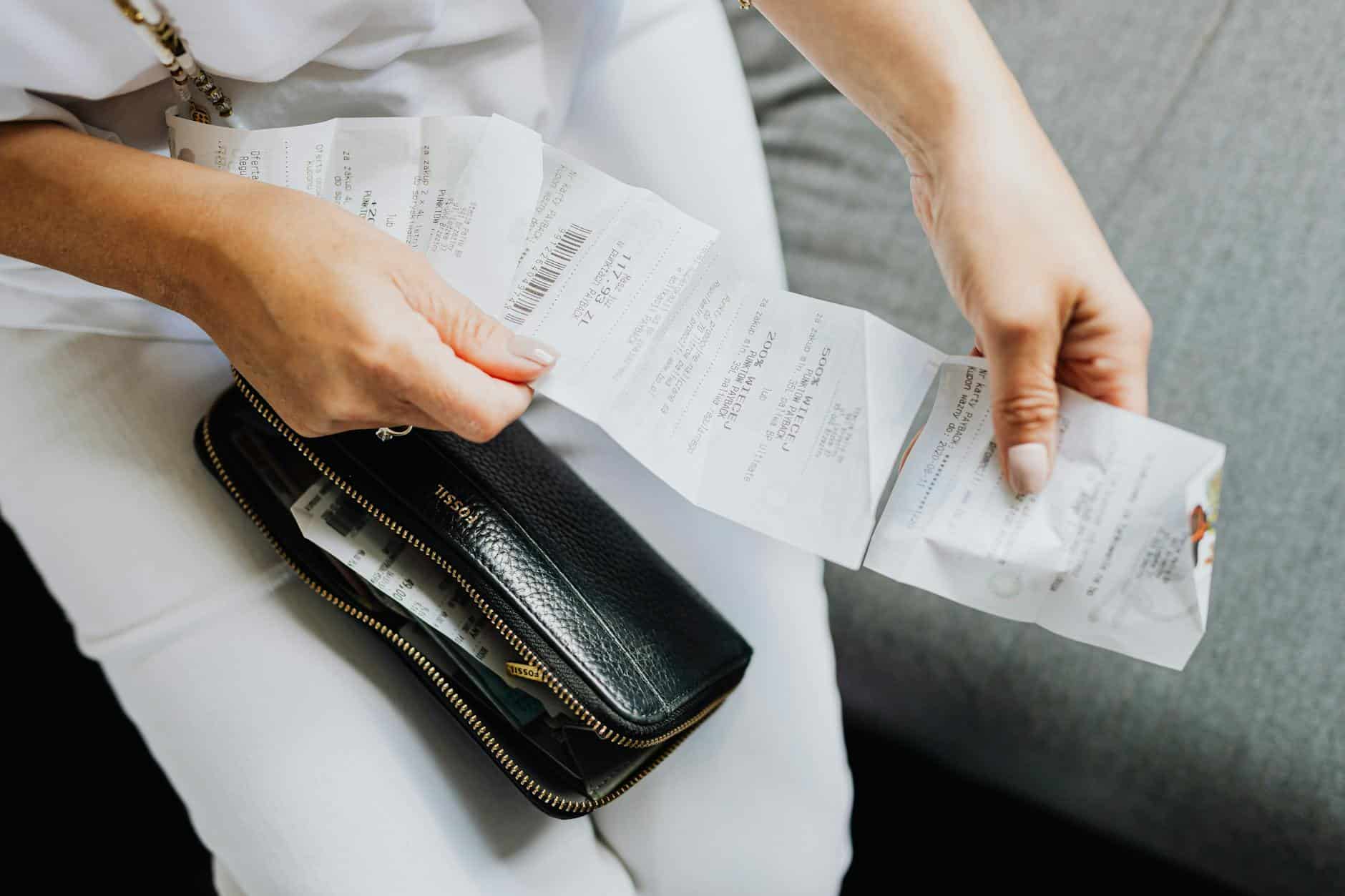 Person in white pants holding receipt papers beside black leather wallet with zipper on gray couch or seat