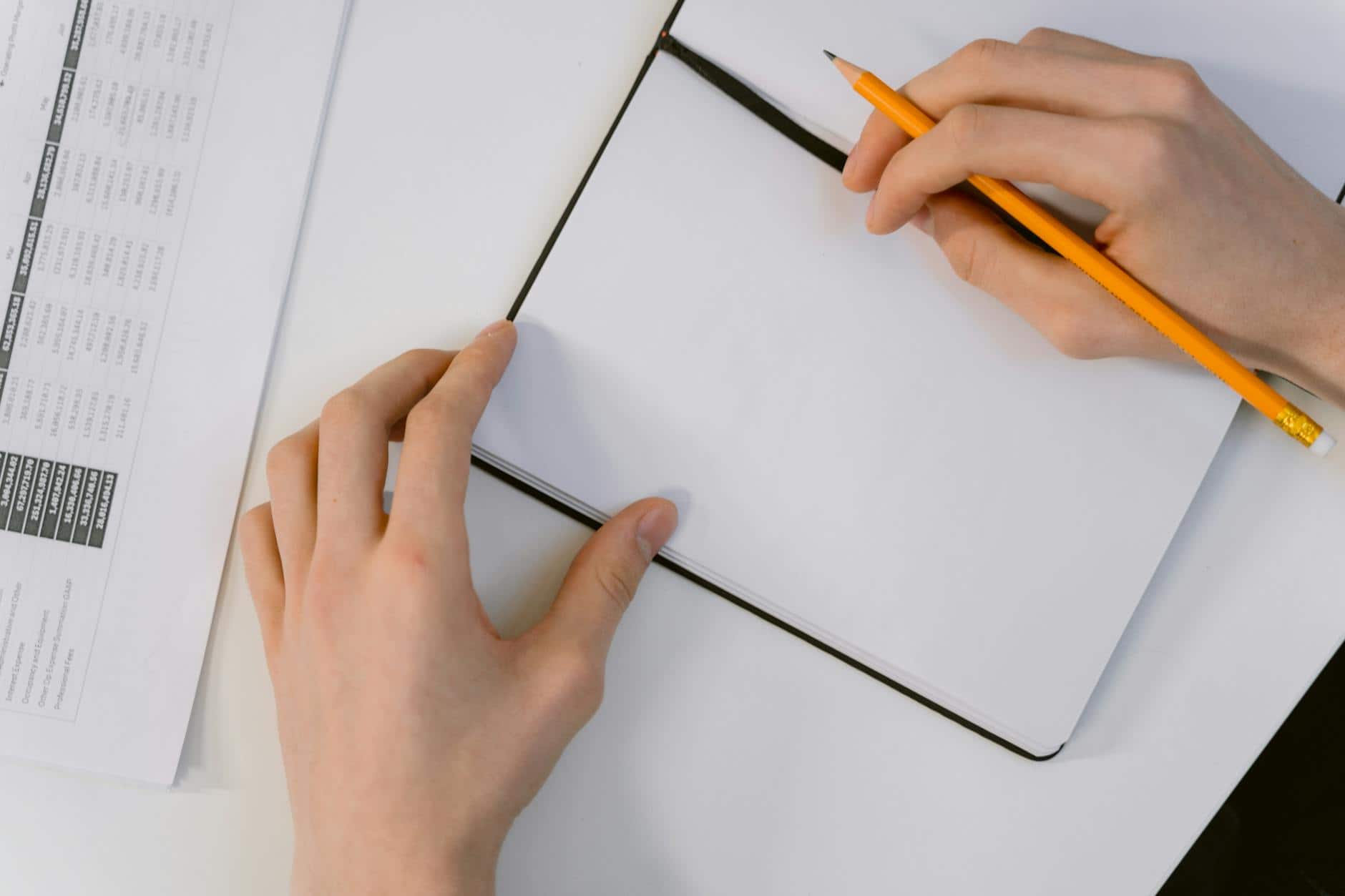 Close-up of hands holding a pencil and writing in a notebook on a white desk.